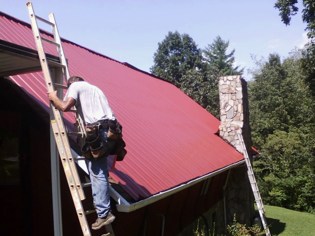 Roofer on a ladder beside a red metal roof and stone chimney — Soffit & Fascia Repair by Roofing Brothers in Harvard
